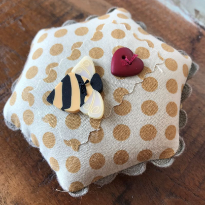 A small fabric pin cushion with a beige and brown polka dot pattern and scalloped edges, decorated with a bumblebee and red heart buttons, resting on a wooden surface.
Photo by Hands on  Design [additional_image_link]