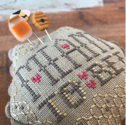 A close-up of a handmade cross-stitch pillow with the word 'MEANT TO BEE' stitched in gray and cream, accented with pink flowers and a honeycomb pattern. Two decorative bees on pins rest on the pillow, which is placed on a wooden surface.
Photo by Hands on  Design [additional_image_link]