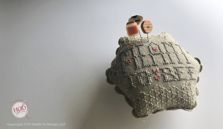 A vintage cross-stitch pincushion in a muted gray-beige fabric with embroidered floral and geometric patterns, featuring two wooden pins with orange and brown tops inserted into the top, set against a plain light gray background.
Photo by Hands on  Design [additional_image_link]