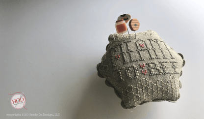 A vintage cross-stitch pincushion in a muted gray-beige fabric with embroidered floral and geometric patterns, featuring two wooden pins with orange and brown tops inserted into the top, set against a plain light gray background.
Photo by Hands on  Design [additional_image_link]