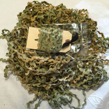 A pile of curly ric rac with a small clear plastic bag containing more of the same and a blank beige tag placed on top, set against a white fabric background. [additional_image_link]