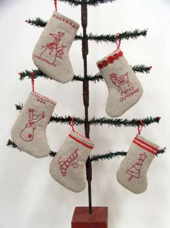 Five beige felt Christmas stockings with red embroidered holiday designs hang from the branches of a dark brown decorative tree against a white background.
Photo by The Birdhouse Patchwork Designs [additional_image_link]