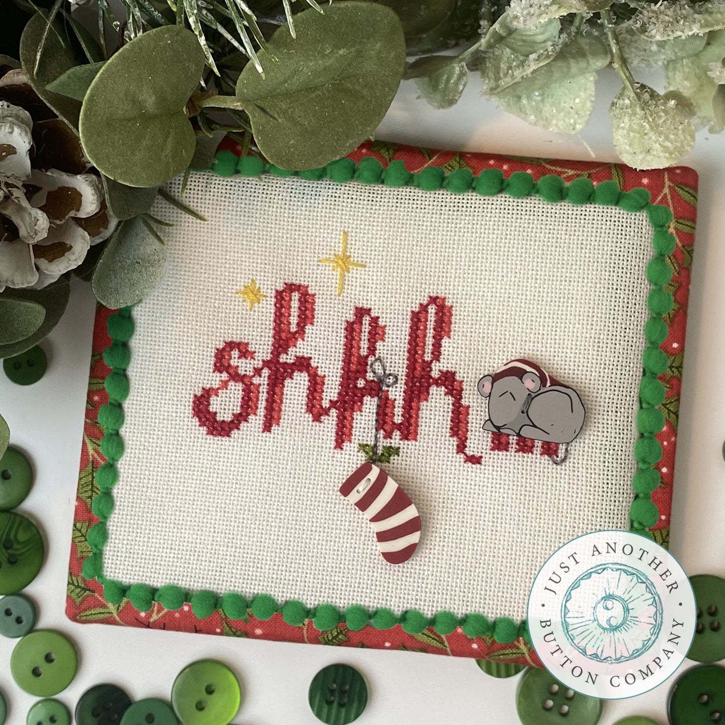 Cross stitch ornament kit depicting a Christmas scene with the text 'SHHH' in red and green, surrounded by festive images including a mouse and holiday decorations on a white background with red and green trimmings.
Photo by JustAnotherButtonCompany.com [image_link]