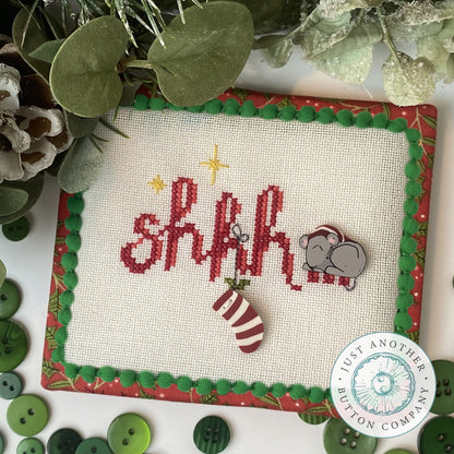 Cross stitch ornament kit depicting a Christmas scene with the text 'SHHH' in red and green, surrounded by festive images including a mouse and holiday decorations on a white background with red and green trimmings.
Photo by JustAnotherButtonCompany.com [image_link]