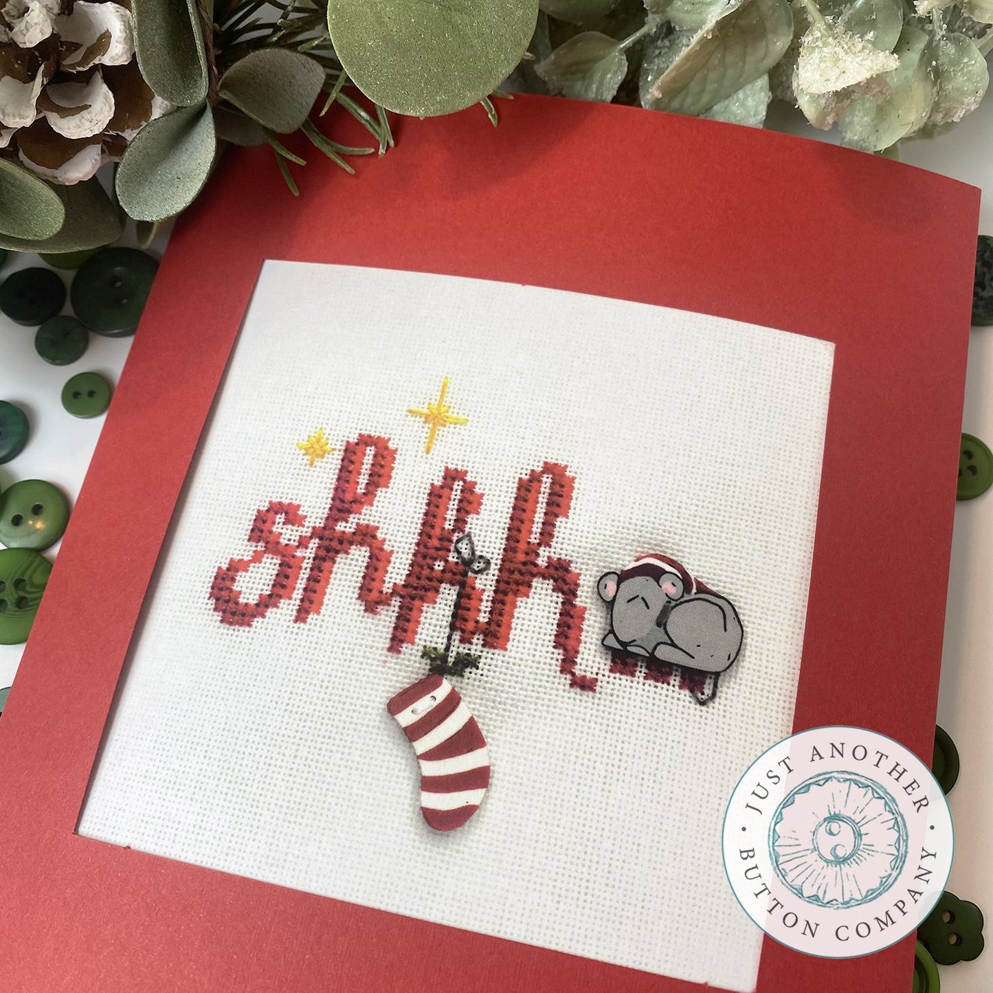 Cross stitch ornament kit depicting a Christmas scene with the text 'SHHH' in red and green, surrounded by festive images including a mouse and holiday decorations on a white background with red and green trimmings.
Photo by JustAnotherButtonCompany.com [additional_image_link]
