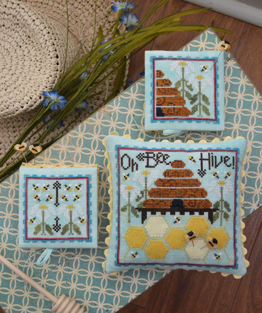A pincushion, needlebook, and needle park cross stitch embroidery kit with bee-themed designs on a seafoam linen background, displayed on a table with a blue-patterned pincushion, a needle park, and floss.