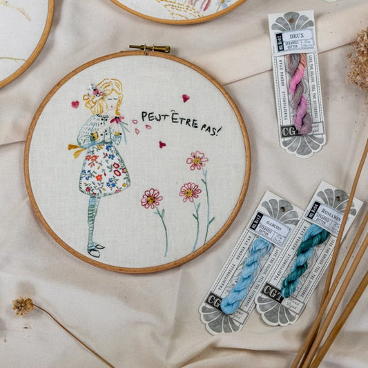 A round embroidery hoop displays a finished floral embroidery of a girl with the French text 'PEUT-ÊTRE PAS!'. Surrounding it are three packages of C6GA brand embroidery floss in pink, blue, and teal, arranged on a beige fabric background with dried flowers and wooden embroidery needles.
Photo by cottagegardenthread.com [image_link]