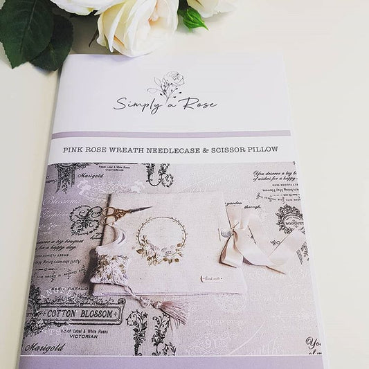 A product cover for a needlecase and scissors pillow pattern by Simply a Rose, featuring a pink rose wreath design on a vintage-style fabric background with a ribbon bow and scissors.
Photo by Angela Watson [image_link]