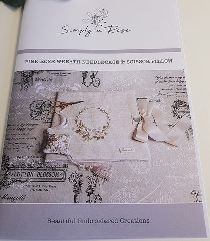 A flat-lay product photo of a 'Simply a Rose' needlecase and scissor pillow kit, titled 'Pink Rose Wreath Needlecase & Scissor Pillow'. The kit is displayed on a white surface with white roses in the top-left corner.
Photo by Angela Watson [additional_image_link]