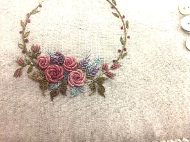 A close-up of a floral embroidery design on a beige linen fabric, featuring pink roses, green leaves, and purple accents.
Photo by Angela Watson [additional_image_link]