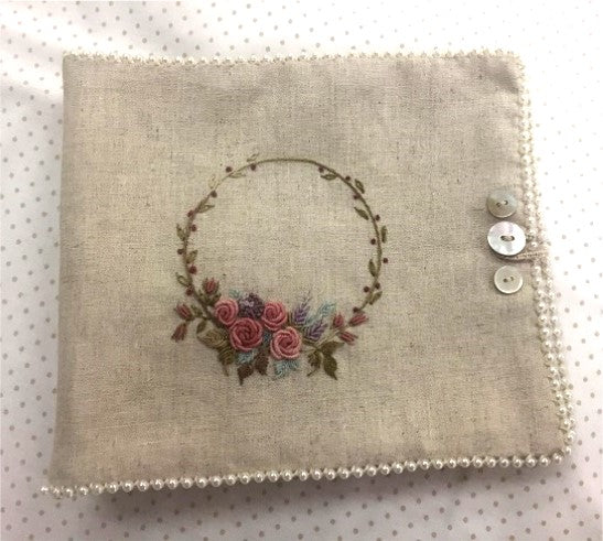 A square beige linen fabric pouch or organizer cover with a floral wreath embroidery in pink, purple, and green, finished with a beaded border and two decorative buttons on the right side, shown on a white polka-dot background.
Photo by Angela Watson [additional_image_link]