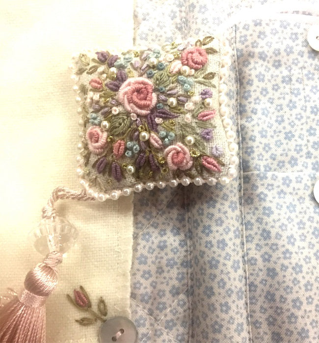 A small embroidered decorative pillow or cushion with pink floral embroidery, pearl-like beads, and a tassel, placed on top of patterned fabric.
Photo by Angela Watson [additional_image_link]