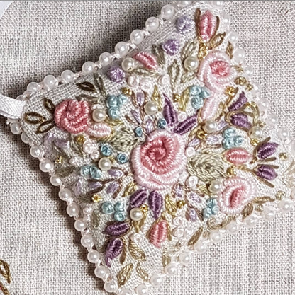 A square embroidered fabric ornament with a floral design, featuring pink and purple roses, green leaves, and white pearl beads along the border, displayed on a textured beige linen background.
Photo by Angela Watson [additional_image_link]