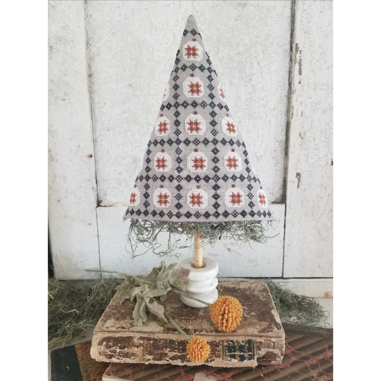 Cross stitch kit depiction: A decorative tree ornament featuring a geometric patterned top with a brown and white color scheme, and a natural-looking tree trunk. Finished product displayed on a rustic background with dried flowers.
Photo by HelloFromLizMathews.com [image_link]