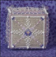 A square, beaded decorative cube with a geometric diamond pattern in beige, white, and blue, set against a purple patterned background.
Photo by JustNan.com [additional_image_link]