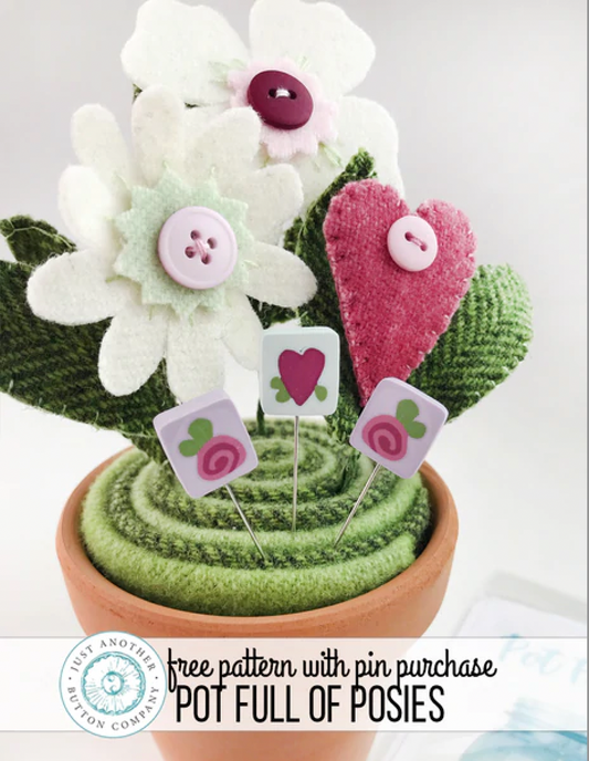 A handmade floral arrangement made from felt flowers, green knitted leaves, and a pink felt heart, displayed in a terracotta pot with decorative pins. A banner at the bottom reads 'free pattern with pin purchase POT FULL OF POSIES' and shows the 'Just Another Button Company' logo.
Photo by JustAnotherButtonCompany.com [image_link]