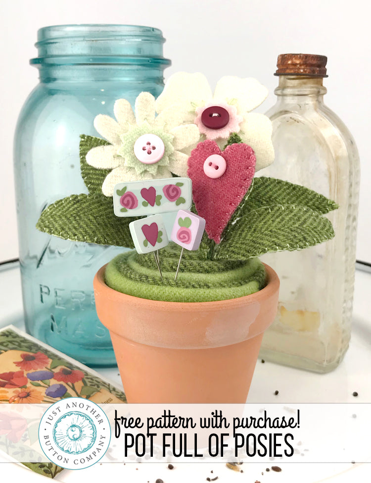 A handmade fabric flower pot arrangement with felt flowers, green textured leaves, and small heart-shaped pins, displayed in a terracotta pot. A blue mason jar and an empty glass bottle stand behind it on a white surface, with a banner at the bottom reading 'free pattern with purchase! POT FULL OF POSIES' and a circular logo for 'The Just Another Company'.
Photo by JustAnotherButtonCompany.com [additional_image_link]