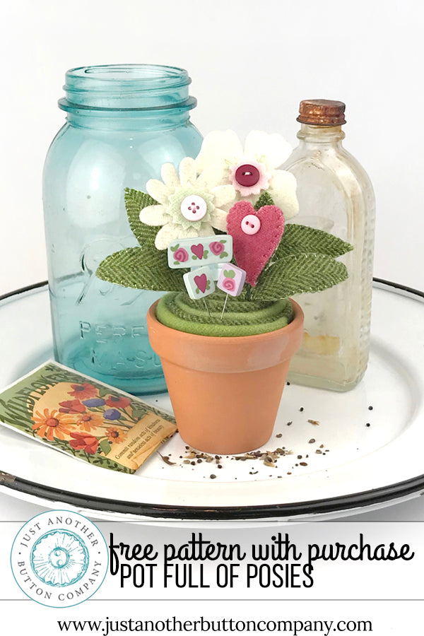 A decorative potted floral arrangement sits on a white tray with a blue glass jar, a clear glass bottle, and a seed packet. The arrangement includes a small terracotta pot with green fabric cactus-like leaves, white felt flowers, a pink felt heart, and small button details.
Photo by JustAnotherButtonCompany.com [additional_image_link]