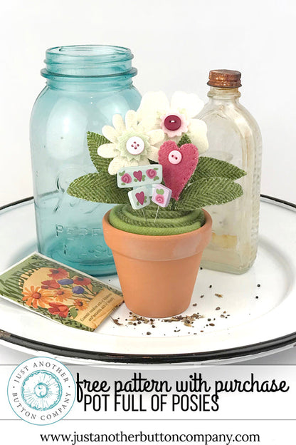 A decorative potted floral arrangement sits on a white tray with a blue glass jar, a clear glass bottle, and a seed packet. The arrangement includes a small terracotta pot with green fabric cactus-like leaves, white felt flowers, a pink felt heart, and small button details.
Photo by JustAnotherButtonCompany.com [additional_image_link]