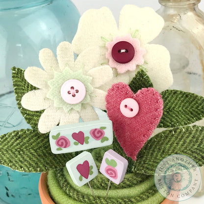 A decorative floral arrangement made from felt and fabric, featuring cream and pink flowers, green leaves, a pink heart-shaped flower, and small white and pink buttons with heart motifs, styled in a pot with glass bottles in the background.
Photo by JustAnotherButtonCompany.com [additional_image_link]