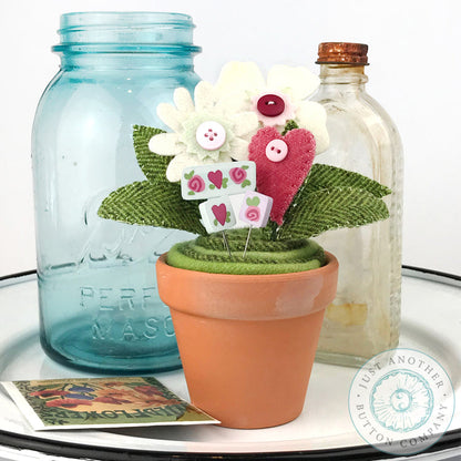 A handmade Valentine's Day felt flower arrangement in a small terracotta pot, styled with a blue mason jar and a glass bottle on a white tray.
Photo by JustAnotherButtonCompany.com [additional_image_link]