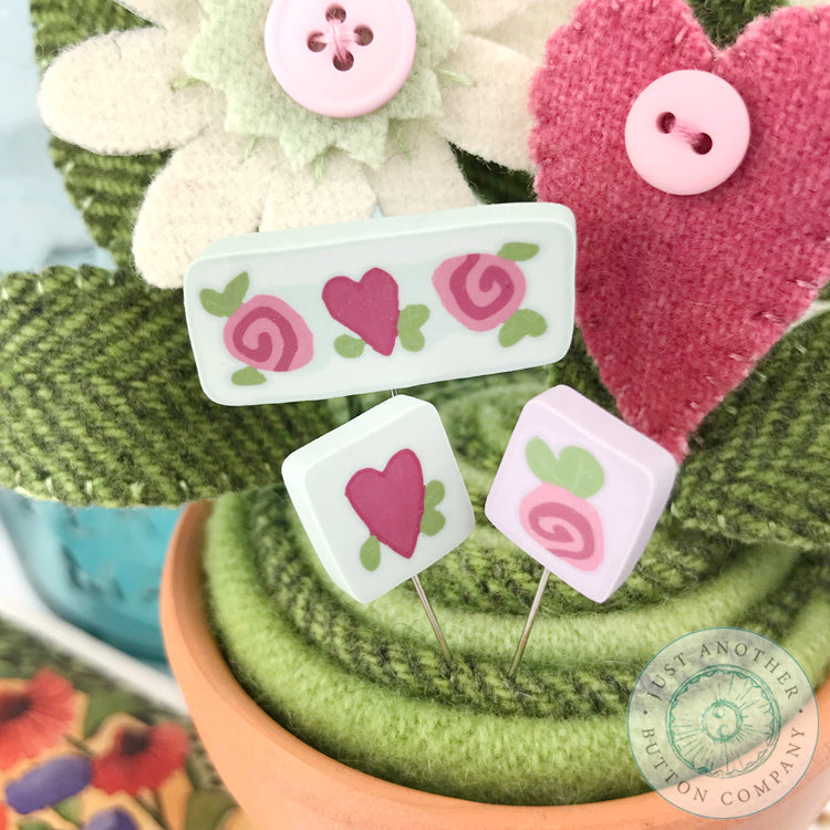 Three small decorative pins with floral and heart motifs displayed in a pot with green fabric leaves and felt flowers. The pins include pastel pink, mint green, and white designs with red/pink hearts and roses.
Photo by JustAnotherButtonCompany.com [additional_image_link]
