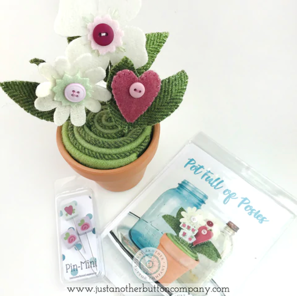 A handmade felt flower arrangement in a small terracotta pot, displayed alongside its packaging. The arrangement features white felt flowers with pink and purple centers, green felt leaves, and a pink felt heart accent.
Photo by JustAnotherButtonCompany.com [additional_image_link]
