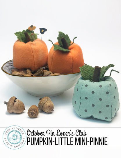 Two mini pumpkin-shaped fabric pins displayed on a white background, one orange with a small acorn and leaf decoration on top, and one light blue with dark blue polka dots. A white bowl with acorns sits behind them, and a banner at the bottom reads 'October Pin Lover's Club PUMPKIN-LITTLE MINI-PINNIE'.
Photo by JustAnotherButtonCompany.com [additional_image_link]