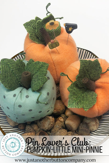 Three mini pumpkin-shaped pin cushions in orange and light blue with green fabric leaves, arranged in a basket with acorns on a white background. A text banner at the bottom reads 'Pin Lover's Club: PUMPKIN-LITTLE MINI-PINNIE' with the website justanotherbuttoncompany.
Photo by JustAnotherButtonCompany.com [additional_image_link]