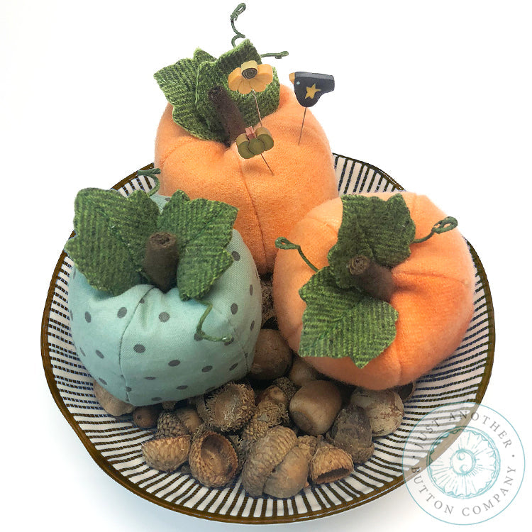 Three decorative fabric pumpkins in orange, sage green, and light blue with polka dots, arranged on a striped plate with acorns, on a white background. A watermark for 'Just Another Button Company' is visible in the lower right corner.
Photo by JustAnotherButtonCompany.com [additional_image_link]
