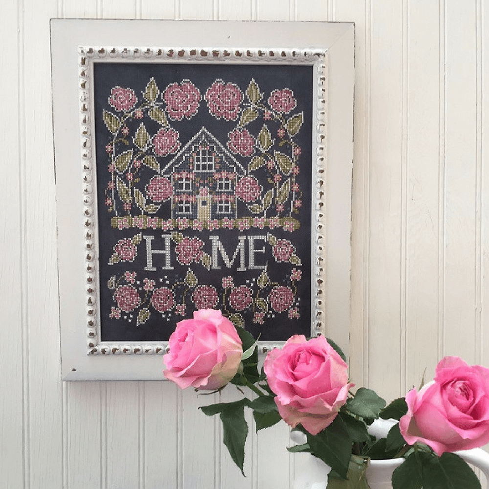 A framed cross-stitch artwork featuring a small house surrounded by pink roses and the word “HOME,” hung on a white paneled wall with pink roses in the foreground.
Photo by HandsOnDesign.biz [image_link]