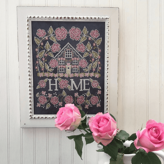 A framed cross-stitch artwork featuring a small house surrounded by pink roses and the word “HOME,” hung on a white paneled wall with pink roses in the foreground.
Photo by HandsOnDesign.biz [image_link]