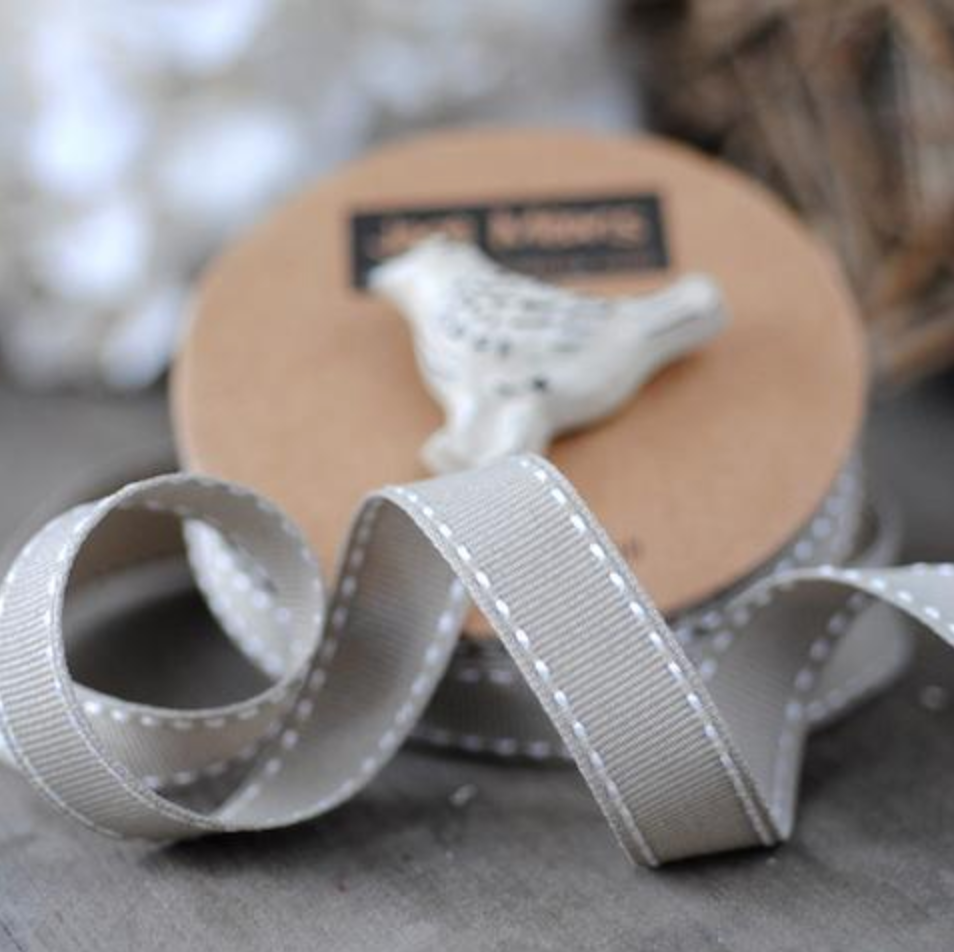 A close-up of a spool of light gray grosgrain ribbon with white dashed stitching, with a small white bird figurine on the cardboard core.
Photo by Janemeans.com [additional_image_link]