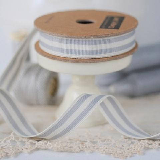 A spool of narrow striped ribbon in light gray and white, displayed on a cream pedestal stand with lace in the foreground and a blurred matching spool in the background.
Photo by Janemeans.com [additional_image_link]