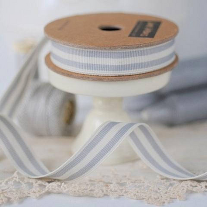 A spool of narrow striped ribbon in light gray and white, displayed on a cream pedestal stand with lace in the foreground and a blurred matching spool in the background.
Photo by Janemeans.com [additional_image_link]