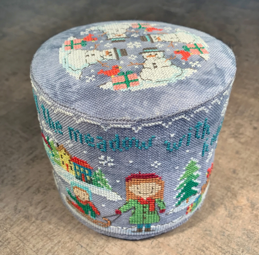 A round, cylindrical needlepoint canvas featuring a winter holiday design with snowmen, snowdrops, a child in a green coat, a house, a Christmas tree, and the text 'the meadow with flowers' on a dusty blue-gray background.
Photo by Tiny Modernist [image_link]