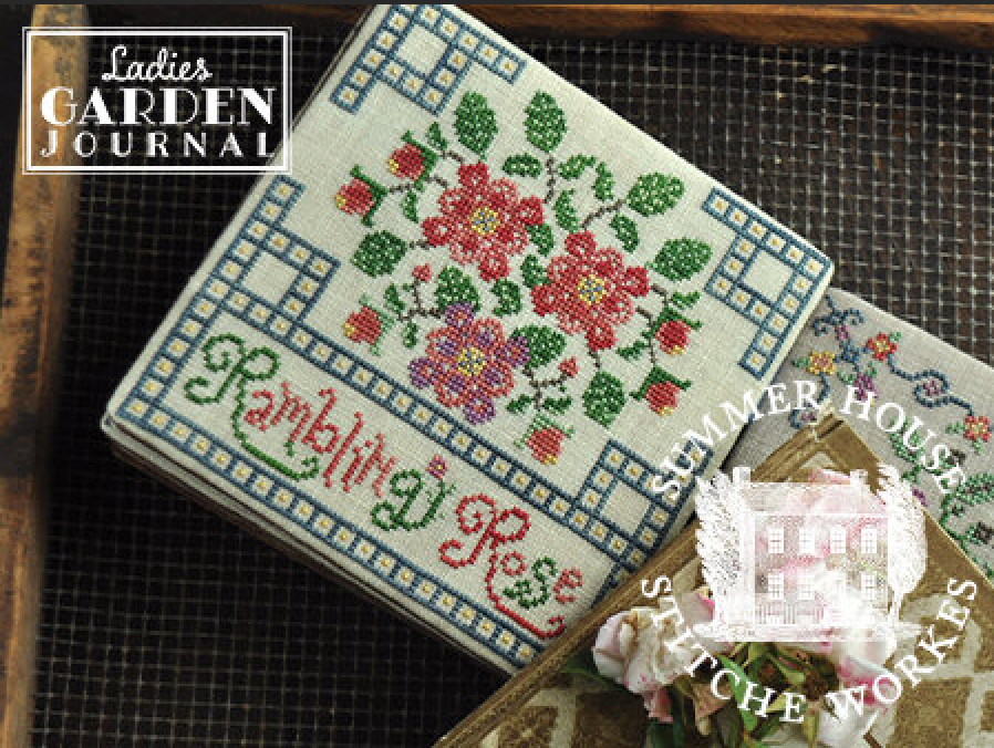 A cross-stitch sampler titled 'Rambling Rose' by Summer House Stitch Works, featuring embroidered pink and red flowers with green leaves on a cream fabric background, styled on a dark mesh tray.
Photo by Summer House Stitche Workes [image_link]