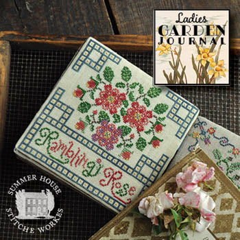 A framed cross-stitch sampler titled 'Bamboo Rose' by Summer House Stitcheworks, displayed on a dark wooden surface next to a 'Ladies Garden Journal' and pink flowers. The sampler features a floral design with pink and red blossoms, green leaves, and a blue geometric border.
Photo by Summer House Stitche Workes [additional_image_link]