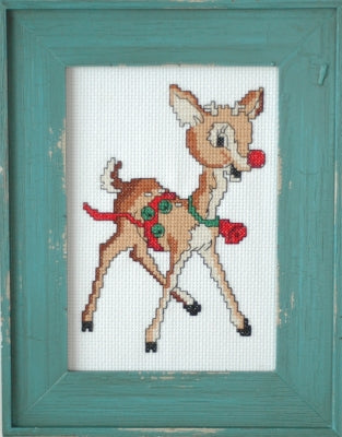 A framed cross-stitch artwork depicting a brown fawn with a red nose and red-and-green holiday harness, set within a distressed teal wooden frame.
Photo by Tiny Modernist [image_link]