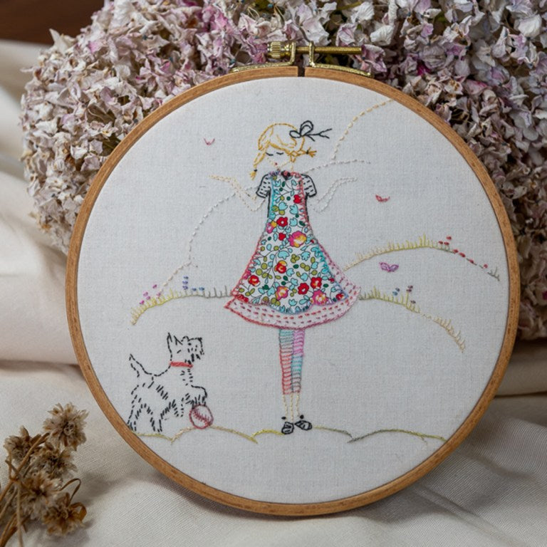 A circular embroidery hoop containing a colorful stitched illustration of a girl in a floral dress with a dog, displayed on a soft fabric background with dried flowers.
Photo by cottagegardenthread.com [image_link]
