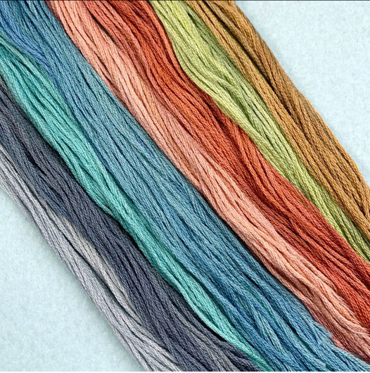 Six skeins of embroidery floss arranged diagonally on a light blue background, showing a gradient of colors from gray and slate blue to teal, mint, coral, orange, and mustard.
Photo by Colour and Cotton [additional_image_link]