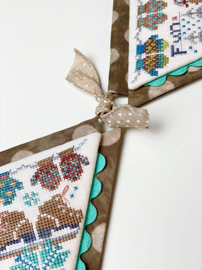 A close-up of a decorative fabric banner with triangular flags tied together by a ribbon. The flags show embroidered winter-themed patterns in brown, teal, blue, and cream, with scalloped brown fabric borders.
Photo by HandsOnDesign.biz [additional_image_link]