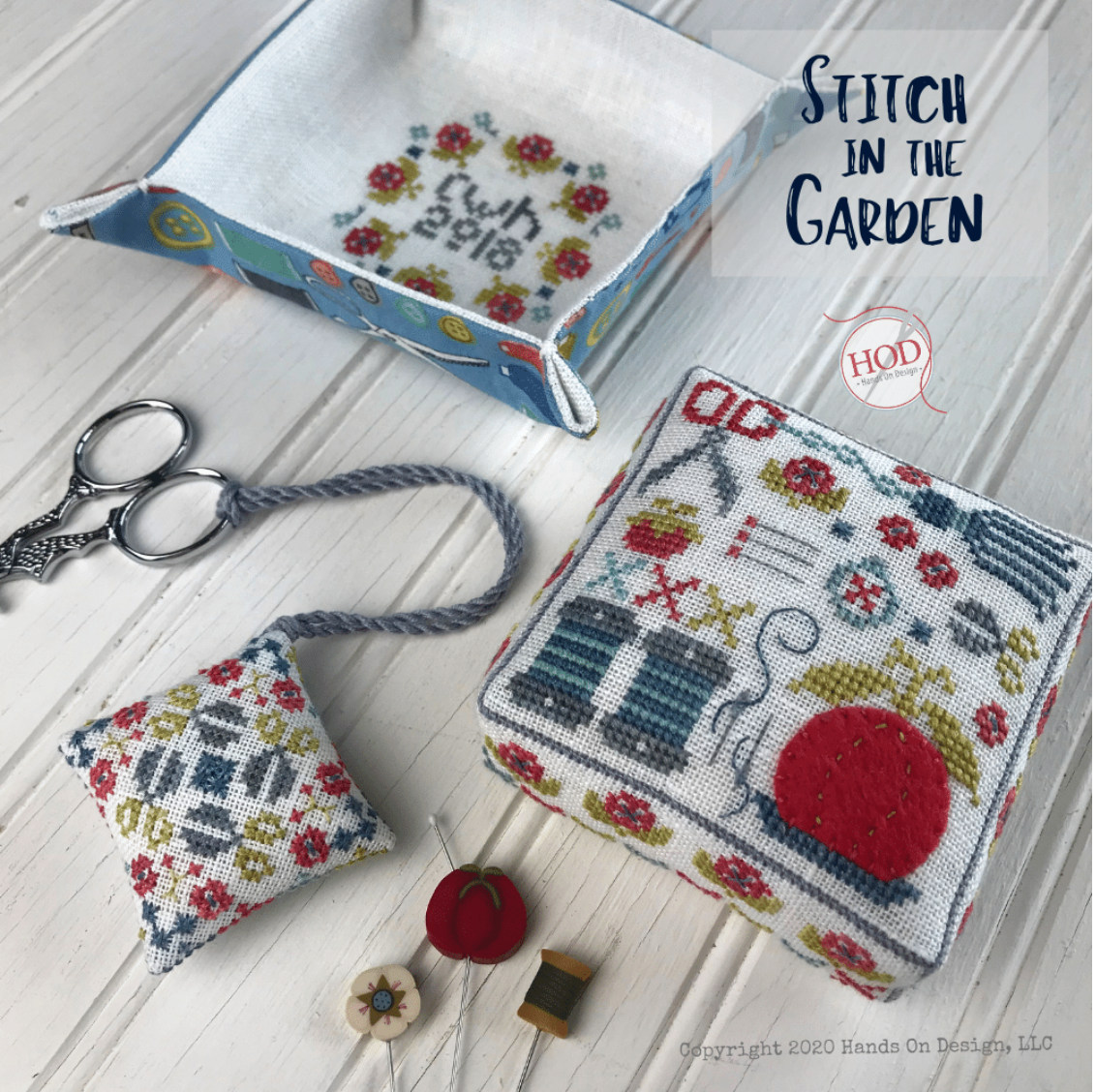 A styled flat-lay of handmade cross-stitch pattern pieces and sewing tools on a white wood background, including a square stitched cushion, a small floral pillow, a fabric tray, scissors, thread, and a spool. Text reads 'Stitch in the Garden' and 'HOD'.
Photo by HandsOnDesign.biz [image_link]