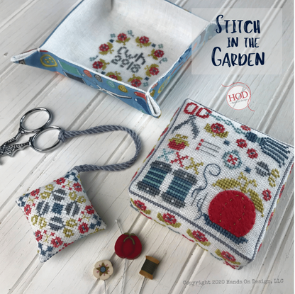 A styled flat-lay of handmade cross-stitch pattern pieces and sewing tools on a white wood background, including a square stitched cushion, a small floral pillow, a fabric tray, scissors, thread, and a spool. Text reads 'Stitch in the Garden' and 'HOD'.
Photo by HandsOnDesign.biz [image_link]