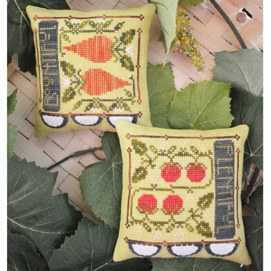 Two square cross-stitch pillows displayed on a woven mat surrounded by green leaves. The top pillow features orange carrots and the bottom pillow features red apples, both with yellow-green backgrounds and dark blue side panels.
Photo by HandsOnDesign.biz [image_link]