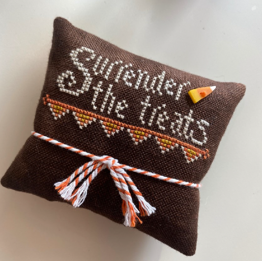 A small brown fabric pillow with white embroidered text reading 'Surrender the treats,' an orange and yellow candy corn accent, and a white-and-orange striped cord tied around the middle, shown on a plain light background.
Photo by HandsOnDesign.biz [image_link]