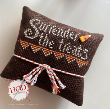 A brown woven fabric pillow with cross-stitched text reading 'Surrender the treats' in white, orange, and yellow. A small candy corn accent is pinned to the top right, and the bottom is tied with an orange and white striped ribbon.
Photo by HandsOnDesign.biz [additional_image_link]