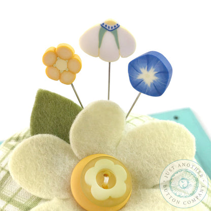 A handmade pincushion kit featuring a floral pattern with yellow wool felt, green wool felt, and a green square print background. Includes the text 'Just Another Button Company' and 'Pin Lover's Club'. Close up of pins on the top.
Photo by JustAnotherButtonCompany.com [additional_image_link]