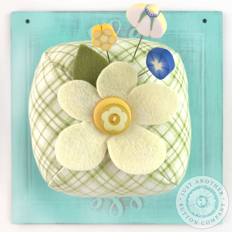 A handmade pincushion kit featuring a floral pattern with yellow wool felt, green wool felt, and a green square print background. Includes the text 'Just Another Button Company' and 'Pin Lover's Club'.
Photo by JustAnotherButtonCompany.com [additional_image_link]