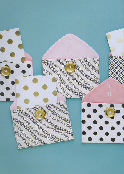 A set of handmade paper envelopes with various patterns including polka dots, stripes, and geometric prints, accented with gold buttons, arranged on a solid teal background.
Photo by See Kate Sew [additional_image_link]